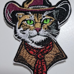 May include: Embroidered patch featuring a cat wearing a cowboy hat and a red neckerchief. The cat has green eyes, a pink nose, and a calico pattern. The hat is black and burgundy. The patch has a white border.