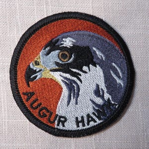 May include: Embroidered circular patch featuring a detailed hawk profile. The design includes a hawk's head with black, white, blue, and yellow details, set against an orange and blue background. The text "AUGUR HAWK" is arched around the bottom.