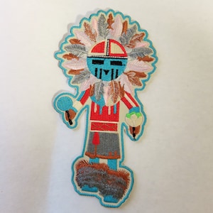May include: Embroidered patch featuring a stylized Native American Kachina doll. The doll has a turquoise face with black markings, a red and white striped torso, and a feathered headdress. It holds a rattle and a bundle. The patch is outlined in turquoise.
