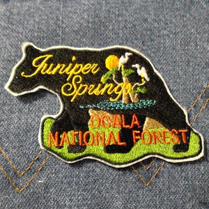 May include: Black embroidered patch shaped like a bear with the text "Juniper Springs Ocala National Forest" in orange and green.