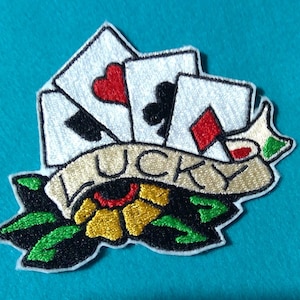 May include: Embroidered iron-on patch with a white background featuring four playing cards, a red heart, a black spade, a black club, and a red diamond. The cards are arranged in a fan shape and are surrounded by a gold banner with the word "LUCKY" in black lettering. The banner is wrapped around a yellow flower with green leaves.