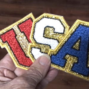 May include: A felt patch with the letters "USA" in red, white, and blue with gold glitter outlining each letter.