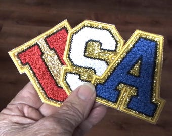 May include: A felt patch with the letters "USA" in red, white, and blue with gold glitter outlining each letter.