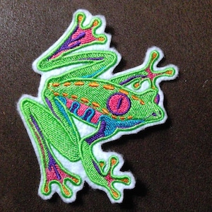 May include: Embroidered patch featuring a vibrant green frog with pink, purple, and blue accents. The frog has a large pink eye and is outlined in bright green stitching. The patch is on a white background.