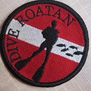 May include: Round embroidered patch with the words "DIVE ROATAN" in black lettering. The patch features a diver silhouette, fish, and a red, white, and black color scheme. The patch is likely for scuba diving enthusiasts.