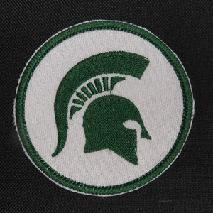 May include: A green and white embroidered patch featuring a Spartan helmet. The helmet is outlined in green and has a white background.