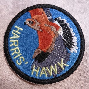 May include: Embroidered patch featuring a Harris' Hawk in flight. The patch is circular with a blue background and a black border. The hawk is depicted in shades of orange, brown, gray, and black, with the words "HARRIS' HAWK" in yellow.