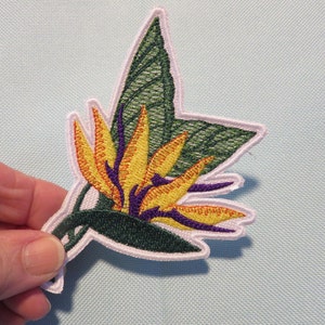 May include: A colorful embroidered patch featuring a bird of paradise flower with yellow, orange, and purple petals and green leaves.