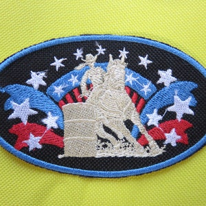 May include: An oval-shaped embroidered patch with a black background. The patch features a white horse and rider, a barrel, and red, white, and blue stars and stripes.