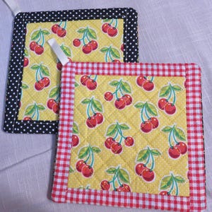 May include: Two quilted pot holders with a yellow cherry pattern. One has a black polka dot border, and the other has a red and white gingham border. Both have a white loop for hanging.