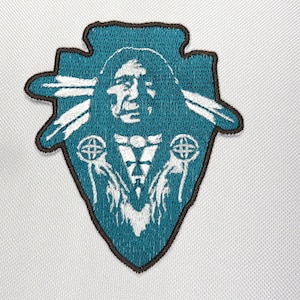 May include: An embroidered patch featuring a Native American chief with a teal background and white accents. The chief is wearing a headdress with feathers and holding two hands up with circles in them.