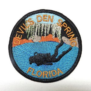 May include: Embroidered patch with a circular design featuring the text "DEVIL'S DEN SPRING FLORIDA". The patch depicts a diver in blue water, a waterfall, and orange and brown rock formations. The border is black.