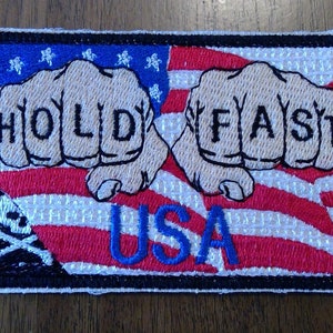 May include: Embroidered patch with a red, white, and blue American flag design. Two fists are shown with the words "HOLD FAST" on the knuckles. The words "USA" are below the fists. A skull and crossbones are in the lower left corner.