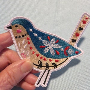 May include: Embroidered iron-on patch of a blue and beige bird with a white flower design. The bird has a long tail and a small beak.