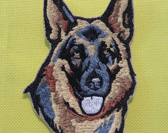 May include: Embroidered patch of a German Shepherd dog's head. The dog has brown and black fur, a black nose, and a white tongue sticking out. The patch is on a green background.