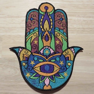 May include: Embroidered Hamsa hand patch with a black border. The design features a central eye, stars, and intricate patterns in teal, blue, orange, purple, green, and gold. This decorative patch is suitable for clothing or accessories.