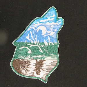 May include: Embroidered patch of a wolf howling at the moon, with a green forest and blue sky in the background. The wolf is outlined in white and the design is on a brown fabric.