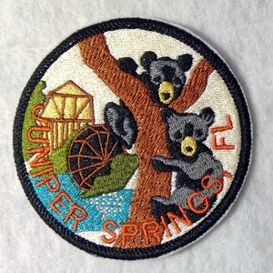May include: Embroidered patch with a white background featuring two black bears climbing a brown tree. The tree is in front of a small wooden building with a water wheel. The text "Juniper Springs FL" is in orange around the edge of the patch.