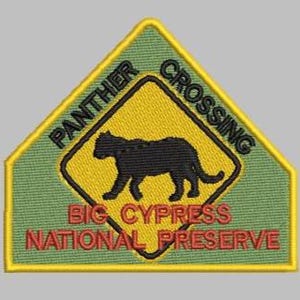 May include: Embroidered patch featuring a black panther silhouette within a yellow diamond, set against a green background. The text "PANTHER CROSSING" arches above, and "BIG CYPRESS NATIONAL PRESERVE" is below. The patch has a yellow border.