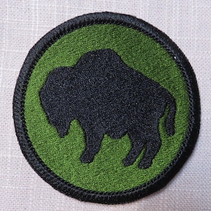 May include: A round patch featuring a black bison silhouette on a green background. The patch has a black border and is likely an emblem or insignia. The design is simple and bold, suitable for various applications.