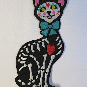 May include: Black and white embroidered patch of a cat skeleton with a red heart and a turquoise bow tie. The cat has a sugar skull face with yellow eyes and pink cheeks.