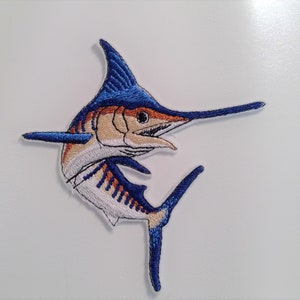 May include: Embroidered patch of a marlin fish in shades of blue, orange, and beige. The fish has a long, pointed bill and a large dorsal fin. The patch is likely for clothing or accessories.