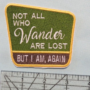 May include: A green and brown embroidered patch with yellow stitching. The patch features the text "NOT ALL WHO Wander ARE LOST BUT I AM, AGAIN".