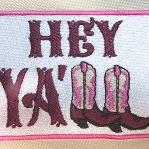 May include: Embroidered patch with the words "HEY Y'ALL" in burgundy script above a pair of pink and white cowboy boots. The patch has a white background with a pink border.