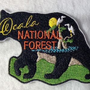 May include: Embroidered patch featuring a black bear silhouette with the text "Ocala National Forest" in orange and yellow. The bear is walking on green grass with a small tree and birds in the background.