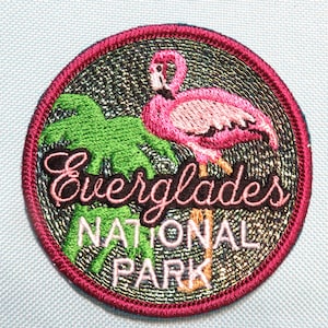 May include: Embroidered circular patch featuring a pink flamingo and green foliage. The patch has the words "Everglades NATIONAL PARK" in a script font. The patch is bordered with a dark pink trim and a metallic background.
