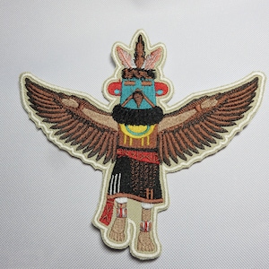May include: Embroidered patch featuring a stylized Native American figure with outstretched wings, wearing a feathered headdress and traditional clothing. The figure is depicted in shades of brown, blue, yellow, and red.