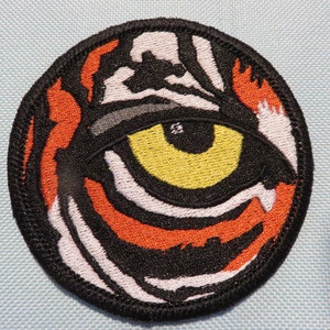 May include: A round embroidered patch with a tiger eye design. The eye is yellow with a black pupil and white sclera. The tiger stripes are orange, black, and white.