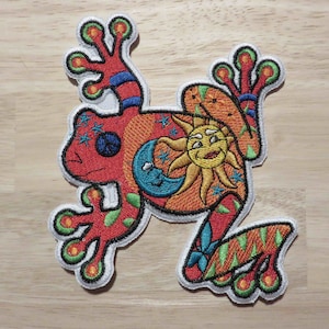 May include: Embroidered iron-on patch featuring a red frog with a sun and moon design. The frog has green, yellow, and blue details.