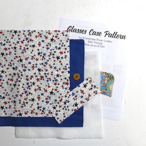 May include: A glasses case pattern with a white fabric featuring red, blue, and black stars. The case has a blue border and a wooden button closure. A printed pattern sheet is also included, with the text "Glasses Case Pattern".