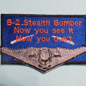 May include: Embroidered patch featuring a silver B-2 Stealth Bomber design over a blue background. The patch includes the text "B-2 Stealth Bomber Now you see it Now you don't" in orange lettering. The patch has a black border.