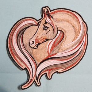 May include: Embroidered patch featuring a horse head within a heart shape. The horse is tan with a flowing mane in shades of red, white, and gray. The patch has a black border and is likely for clothing or accessories.