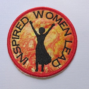 May include: A red and white circular patch with a silhouette of a person with arms raised in the air. The text "INSPIRED WOMEN LEAD" is written around the edge of the patch.