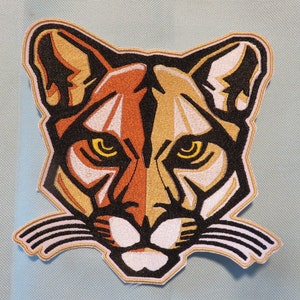 May include: Embroidered patch featuring a stylized cougar face. The design uses orange, gold, black, and white thread to create a geometric representation of the animal's features. The patch has a white border and is likely for application on clothing or accessories.