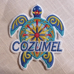 May include: Embroidered patch in the shape of a sea turtle, featuring the word "COZUMEL" in blue. The turtle design includes a compass rose and decorative elements in shades of blue, yellow, red, and gold, with a white border.