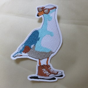 May include: Embroidered patch of a blue and white seagull wearing brown sneakers and sunglasses. The seagull is standing on a white background.