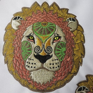 May include: Embroidered lion face with a detailed design. The lion's mane is a mix of gold and brown, with a pink and brown face. The eyes are yellow, and the face has green and brown accents. The design is on a white background.