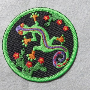 May include: A green embroidered patch with a black background featuring a green lizard with a purple and red stripe down its back. The lizard is surrounded by red and green flowers.
