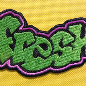 May include: Green embroidered patch with the word "Rock" in a graffiti style font. The patch has a pink outline and is sewn onto a yellow fabric.