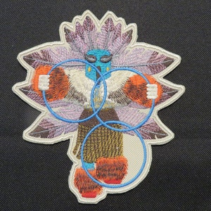 May include: Embroidered patch featuring a colorful, stylized figure with feathers, holding two blue hoops. The figure has a blue face with white eyes and a brown body with a brown skirt.