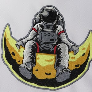 May include: Embroidered patch featuring an astronaut seated on a yellow crescent moon. The astronaut is wearing a gray spacesuit with red accents. The moon has a dark border and a smiling face.