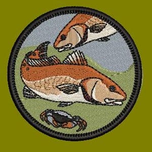 May include: An embroidered patch featuring two redfish and a crab on a green background. The patch is circular and has a black border.