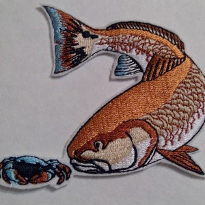 May include: Embroidered patch featuring a redfish with a blue and brown crab. The redfish is orange and white with blue fins. The crab is blue and brown with white claws.