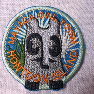 May include: Embroidered patch featuring a gray owl totem design with black accents, surrounded by a blue border. The patch includes text that reads "Hontoon Island Maya Owl Totem". The patch has a green border.