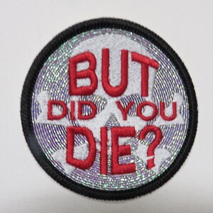May include: Round embroidered patch with the text "BUT DID YOU DIE?" in red letters. The patch has a black border and a silver and purple background. The patch is circular.
