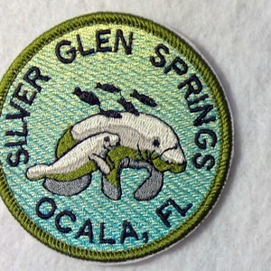 May include: Embroidered patch with a green border, depicting two manatees swimming in blue water with the text "SILVER GLEN SPRINGS OCALA, FL" around the edge.